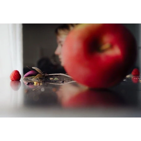 Close-up of a glossy black table with an apple and a raspberry in focus, a blurred profile of a fair-skinned human in background.