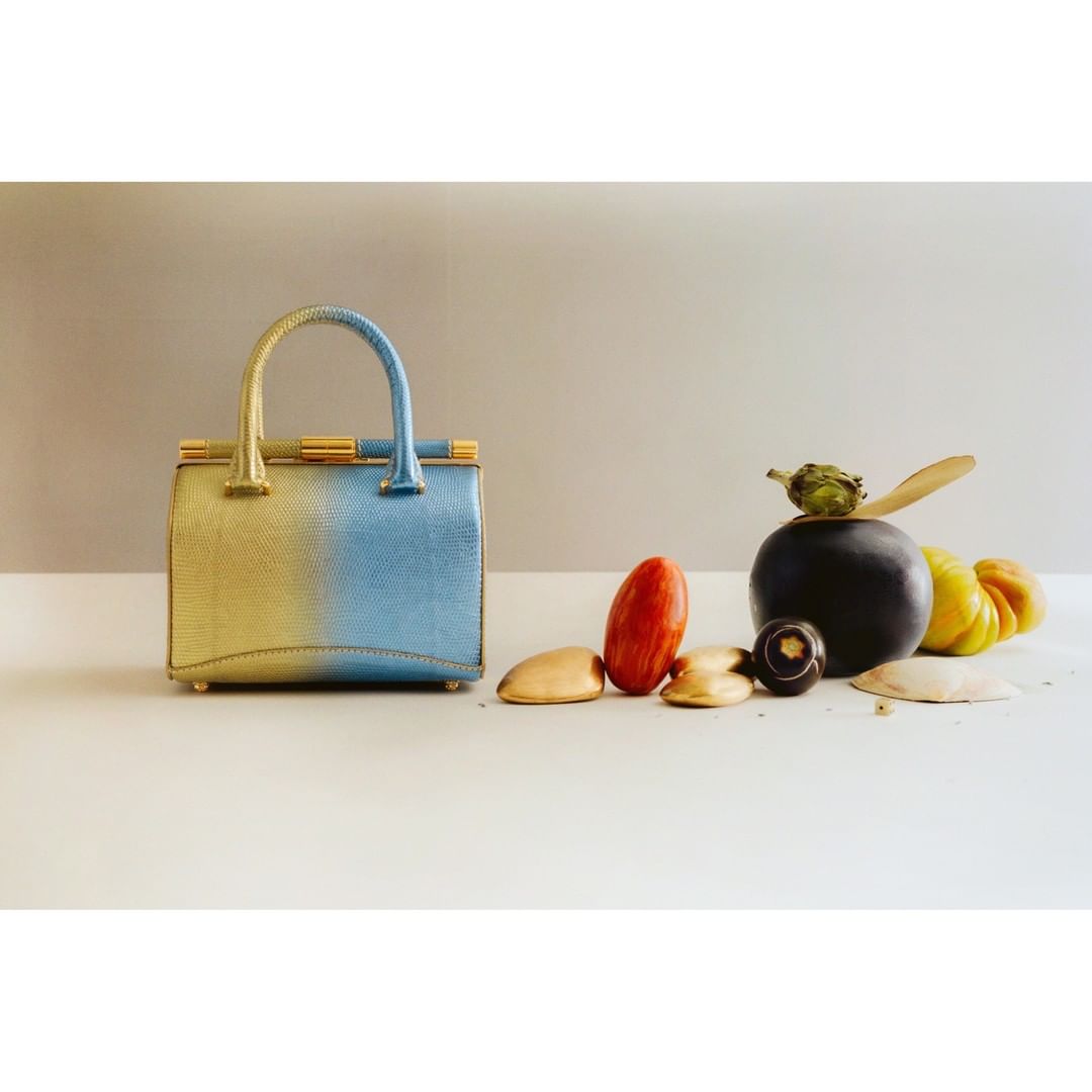 Luxury handbag in two-tone gold and blue, surrounded by a collection of gourds and decorative objects on a neutral background.