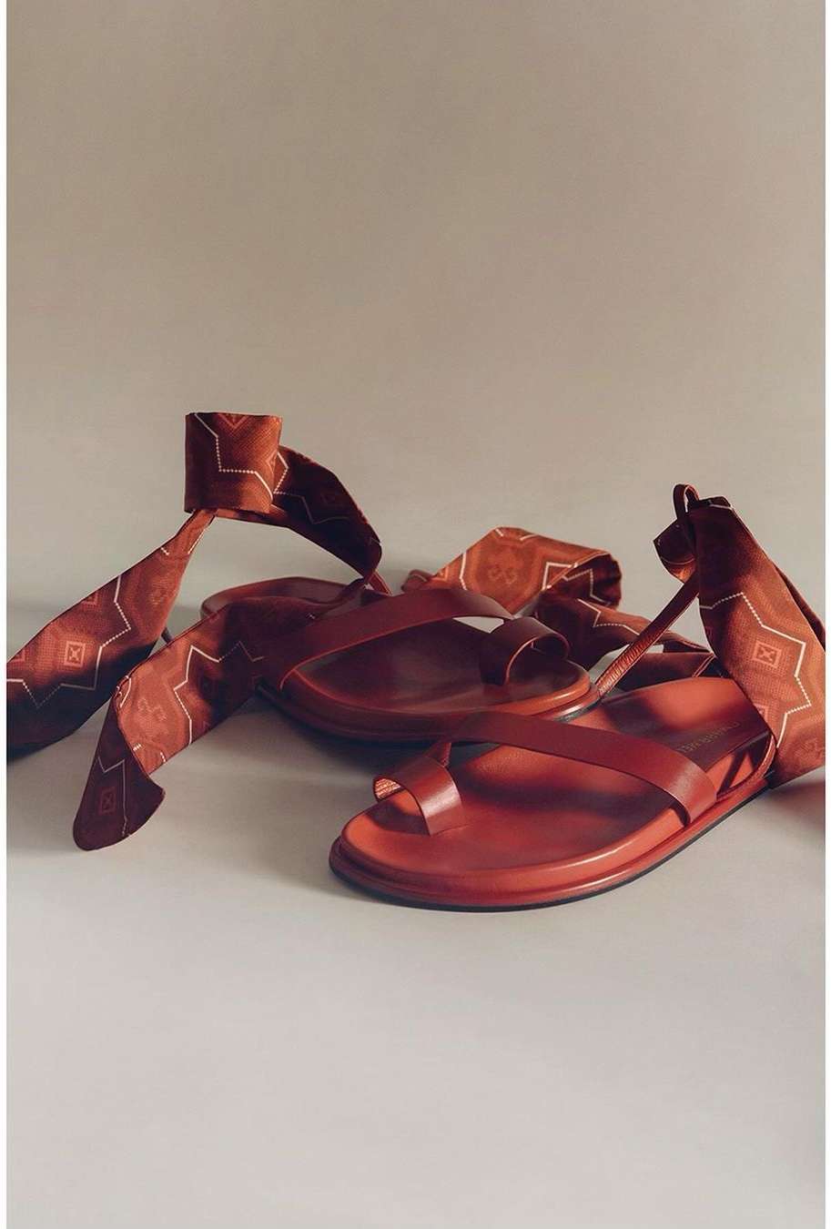 Two reddish-brown leather sandals with toe loops and ankle ties with patterned ribbons on a light grey surface in a studio setting.