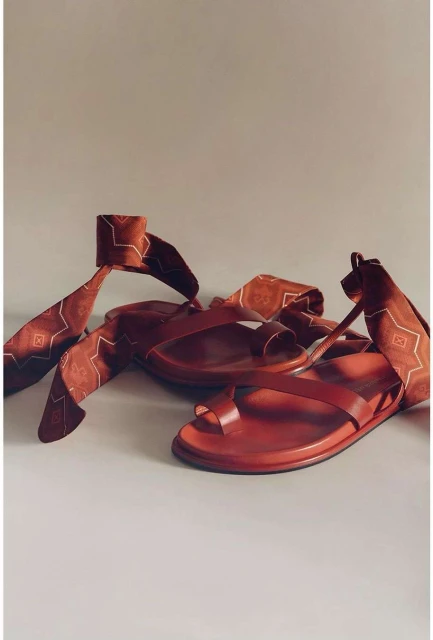 Two reddish-brown leather sandals with toe loops and ankle ties with patterned ribbons on a light grey surface in a studio setting.