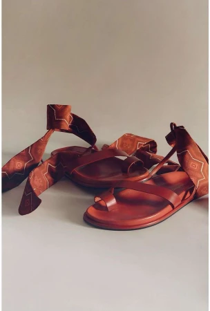 Two reddish-brown leather sandals with toe loops and ankle ties with patterned ribbons on a light grey surface in a studio setting.