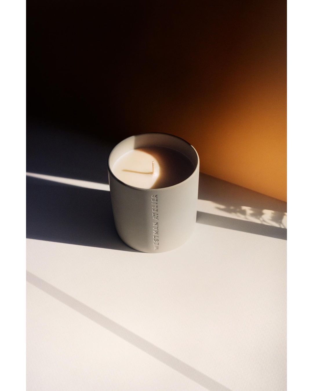 Minimalist product shot of a WESTMAN ATELIER candle in a gray ceramic container, casting soft shadows on a white surface with an orange backdrop.