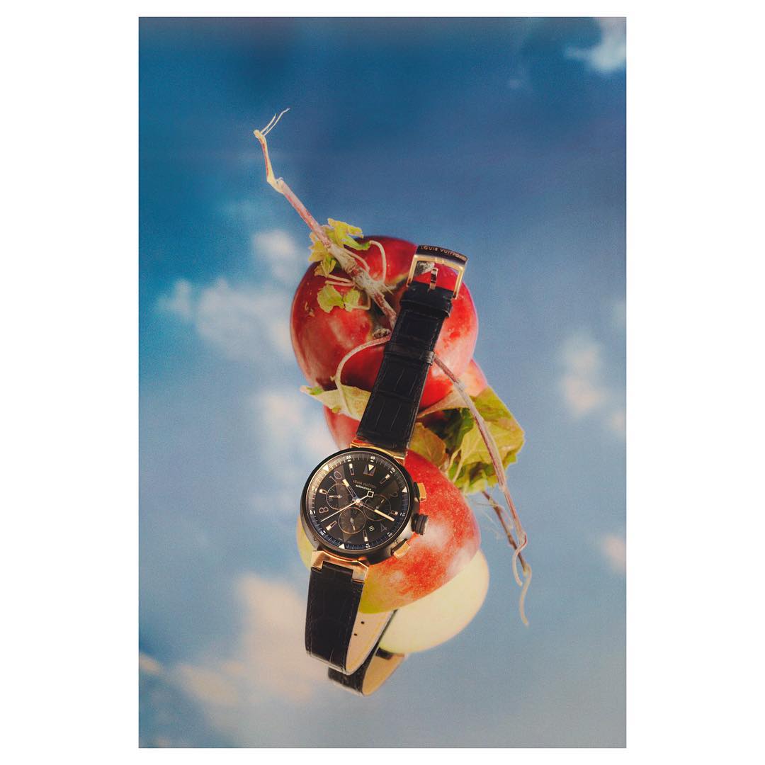 Still life shot featuring a gold wristwatch with a black band on a stack of red apples against a sky-blue background with clouds.