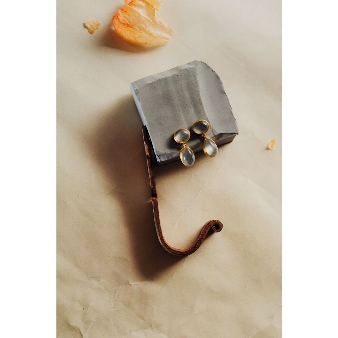 Elegant teardrop earrings in pale grey with gold trim sit on a grey platform with tan textured surface and a peach petal.