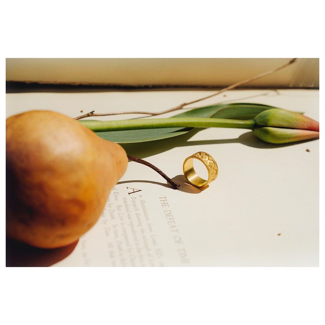 A close-up of a gold ring and a green tulip stem laid on a book page with a brown pear on the side, lit by natural light.