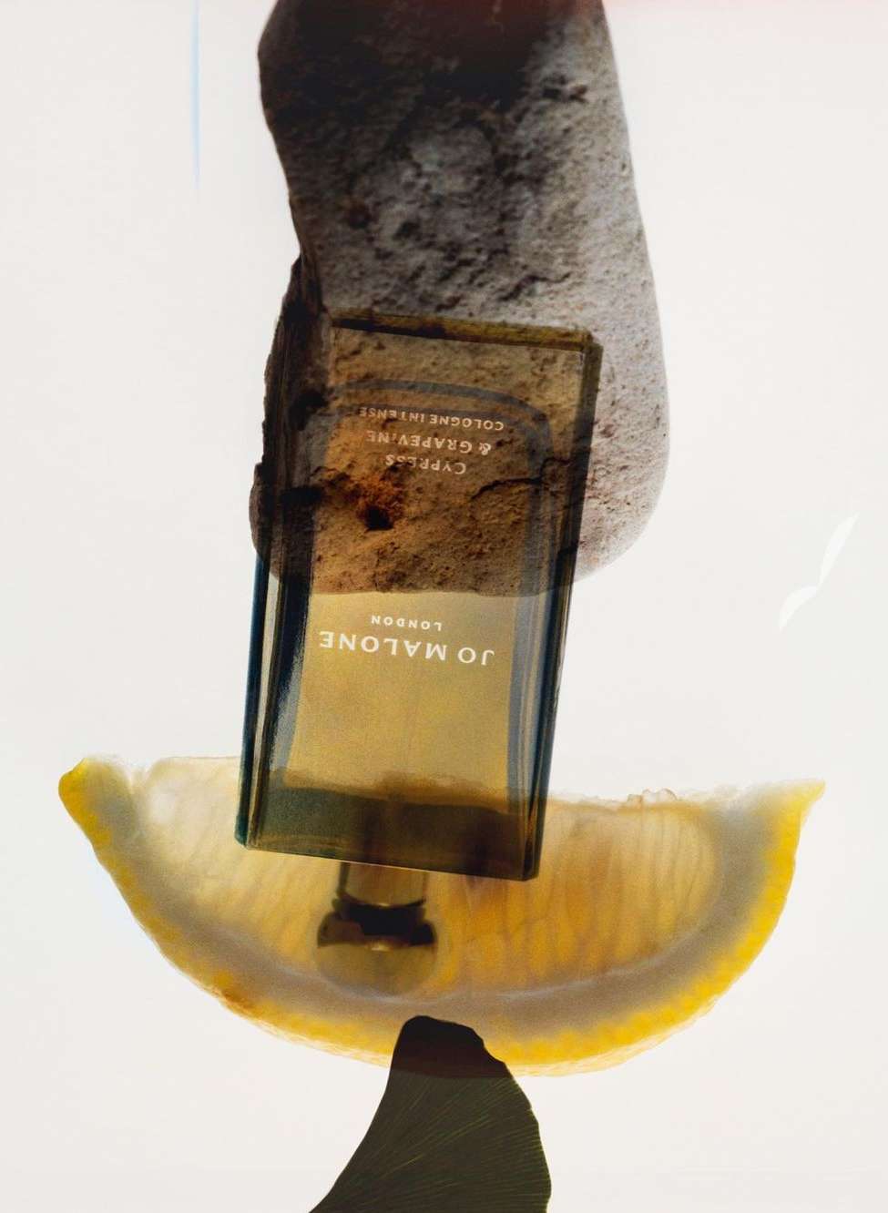 Still life featuring a Jo Malone London cologne bottle balanced on a lemon slice and a rock against a white backdrop.