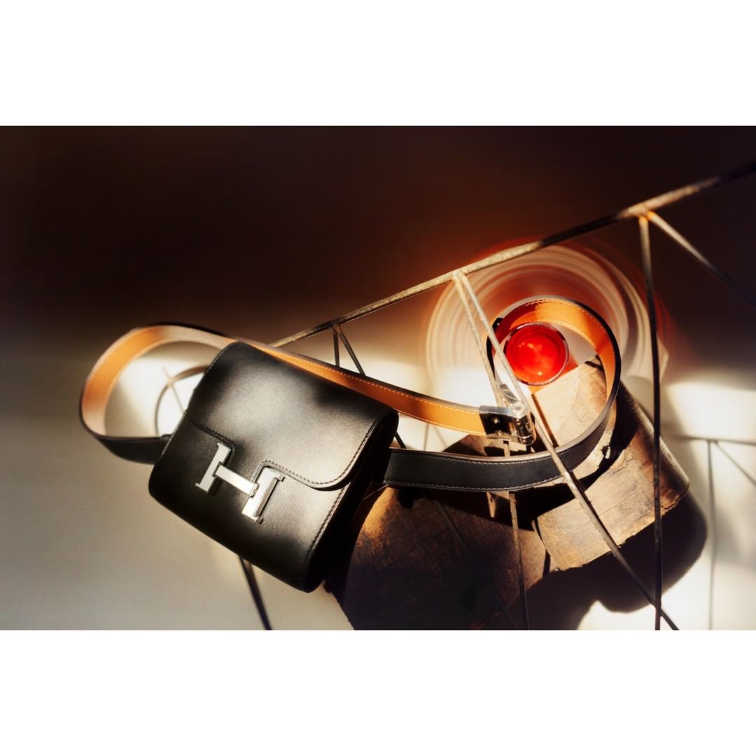 Elegant black leather handbag with a silver logo on a light surface with geometric shadows and shapes for a modern luxury fashion advertisement.