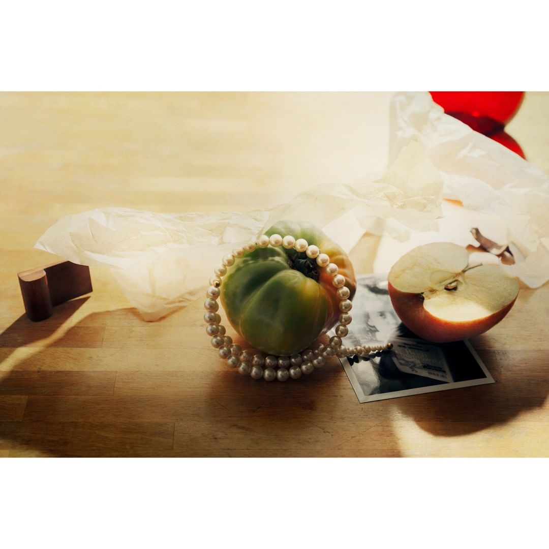Still life featuring a green heirloom tomato adorned with a pearl necklace, alongside a halved apple and a vintage photograph on wood.
