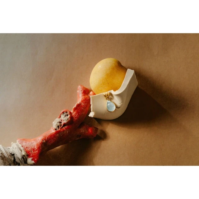 Still life shot of a yellow pear resting in a white plaster container with a red coral branch and a blue pendant on a beige background.