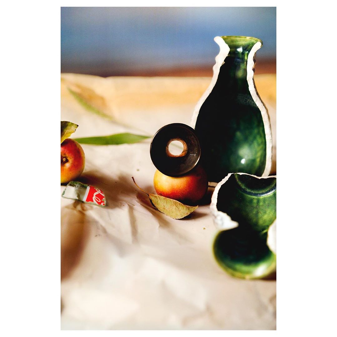 Still life photography featuring a partially broken green ceramic vase, a black ring, red apples, and leaves on a wrinkled paper surface.