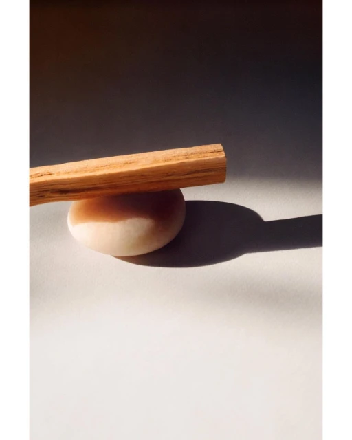 Palo santo stick resting on a round stone against a grey and white background, illuminated by warm, natural lighting.