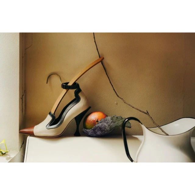 Elegant women's high heel shoe with ankle strap displayed with a wooden hanger, mango fruit and white pitcher on a white surface.