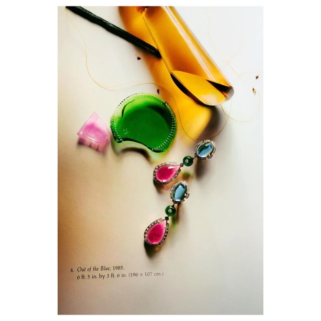 Still life product shot of jewel earrings with pink and blue gemstones, placed alongside colorful glass objects and a flower stem.