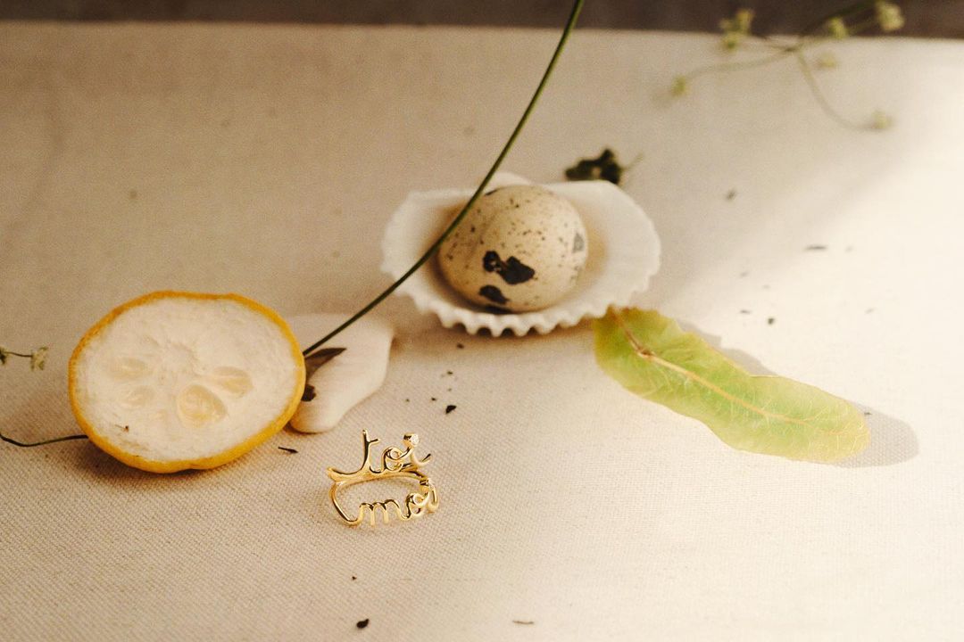Overhead shot of a gold ring spelling 'toi et moi' with a lemon slice, speckled egg in a shell, and green leaf on beige surface.