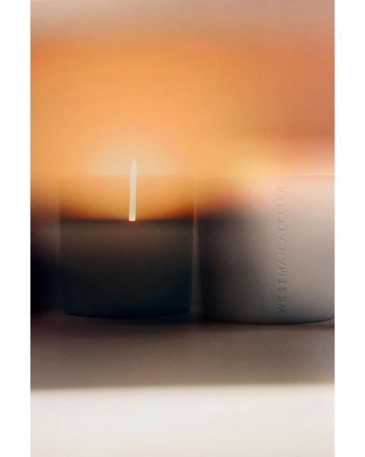 Close-up studio shot of a candle emitting light with the brand Westman Atelier visible on the side of the candle container.