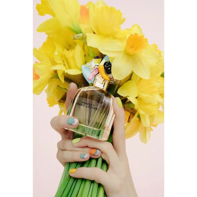 Close-up studio shot of a 'Perfect Marc Jacobs' perfume bottle held by a woman with floral-patterned nails, set against a bouquet of daffodils.