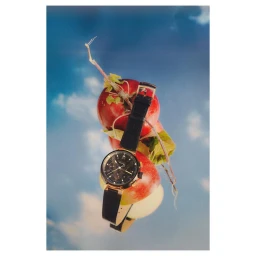 A black leather chronograph watch resting vertically against red apples and green stems against a blue sky backdrop