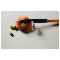 A knife with a yellow handle rests across a peach alongside two small yellow dice and a green berry.