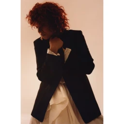 A woman with curly red hair wearing a dark blazer over a white shirt against a warm neutral background