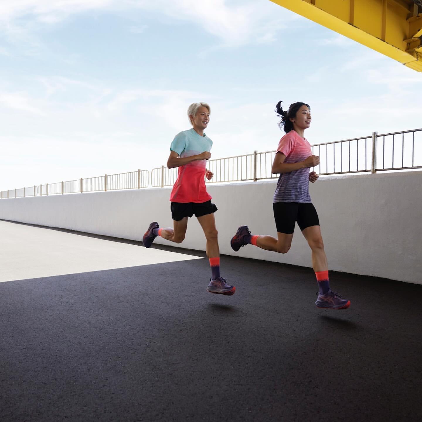 Two young adult female runners stride confidently on a bridge in athletic wear, with blue sky and a yellow structure overhead.