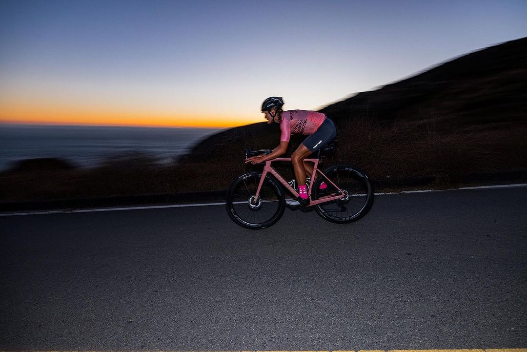 Adult male cyclist in pink jersey rides pink road bike on asphalt road at dusk near the coastline with the sun setting behind him.