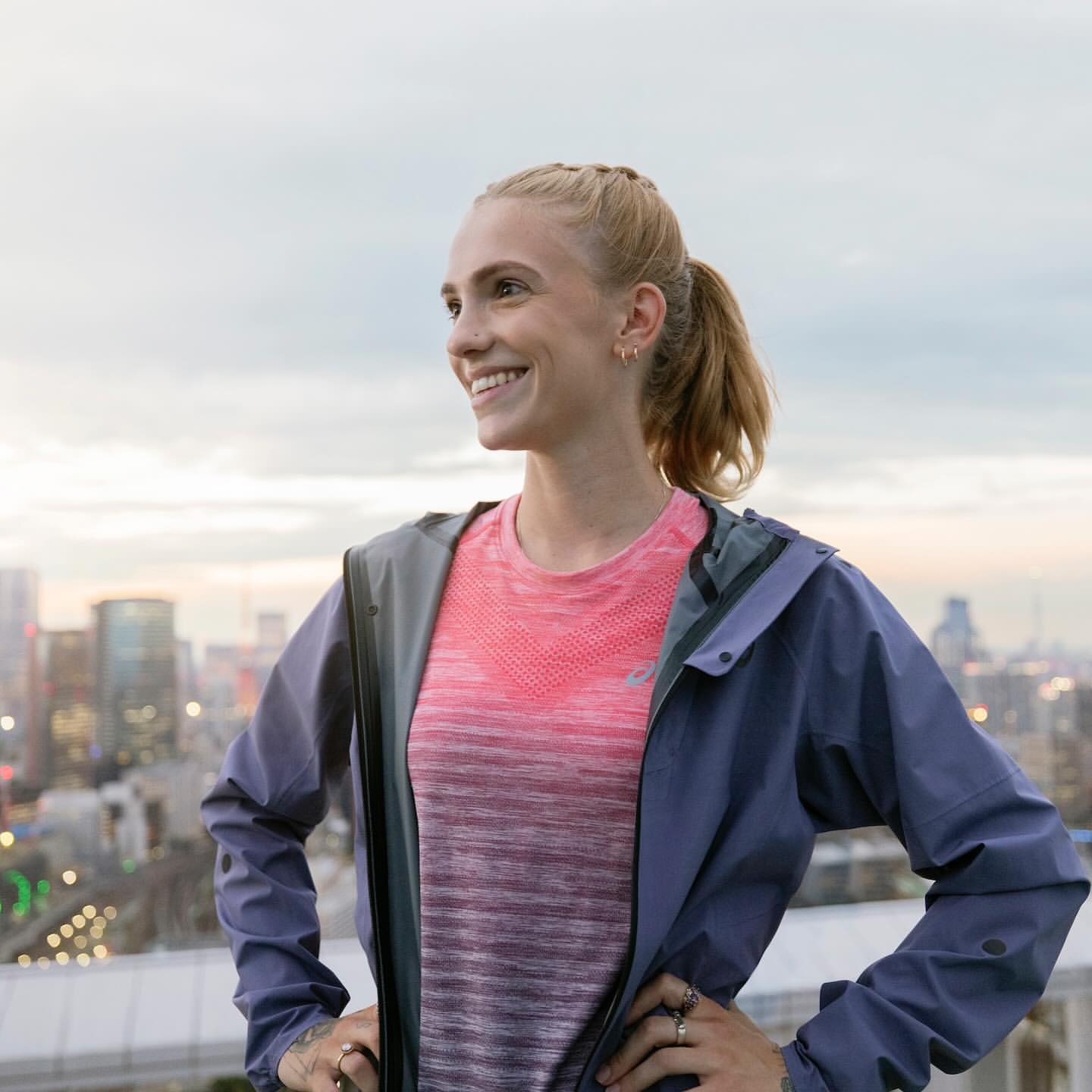Smiling woman in athletic wear stands on a rooftop with a city skyline in the background during daytime, looking off to the side.
