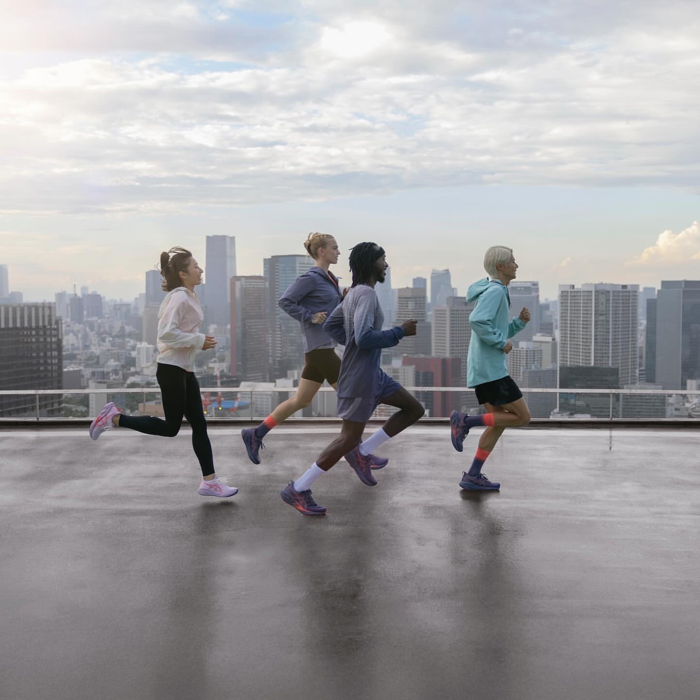 Four athletic women run on a rooftop overlooking a cityscape. They are wearing athletic wear and running shoes during a workout.