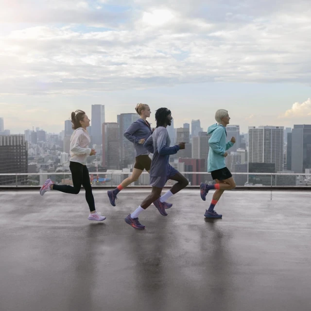 Four athletic women run on a rooftop overlooking a cityscape. They are wearing athletic wear and running shoes during a workout.