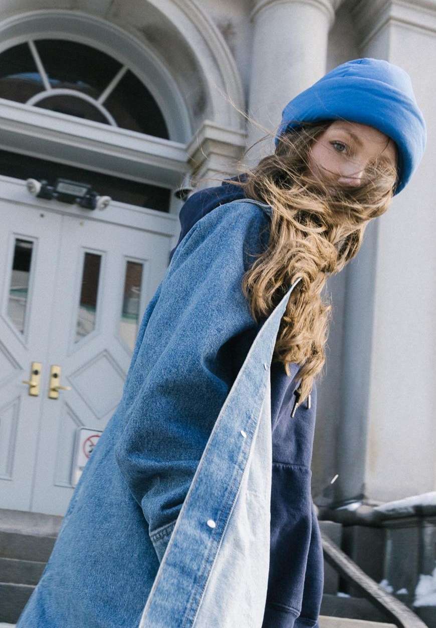 Woman with windswept hair wearing a blue beanie and a two-toned denim and navy jacket, posing outdoors near a building.