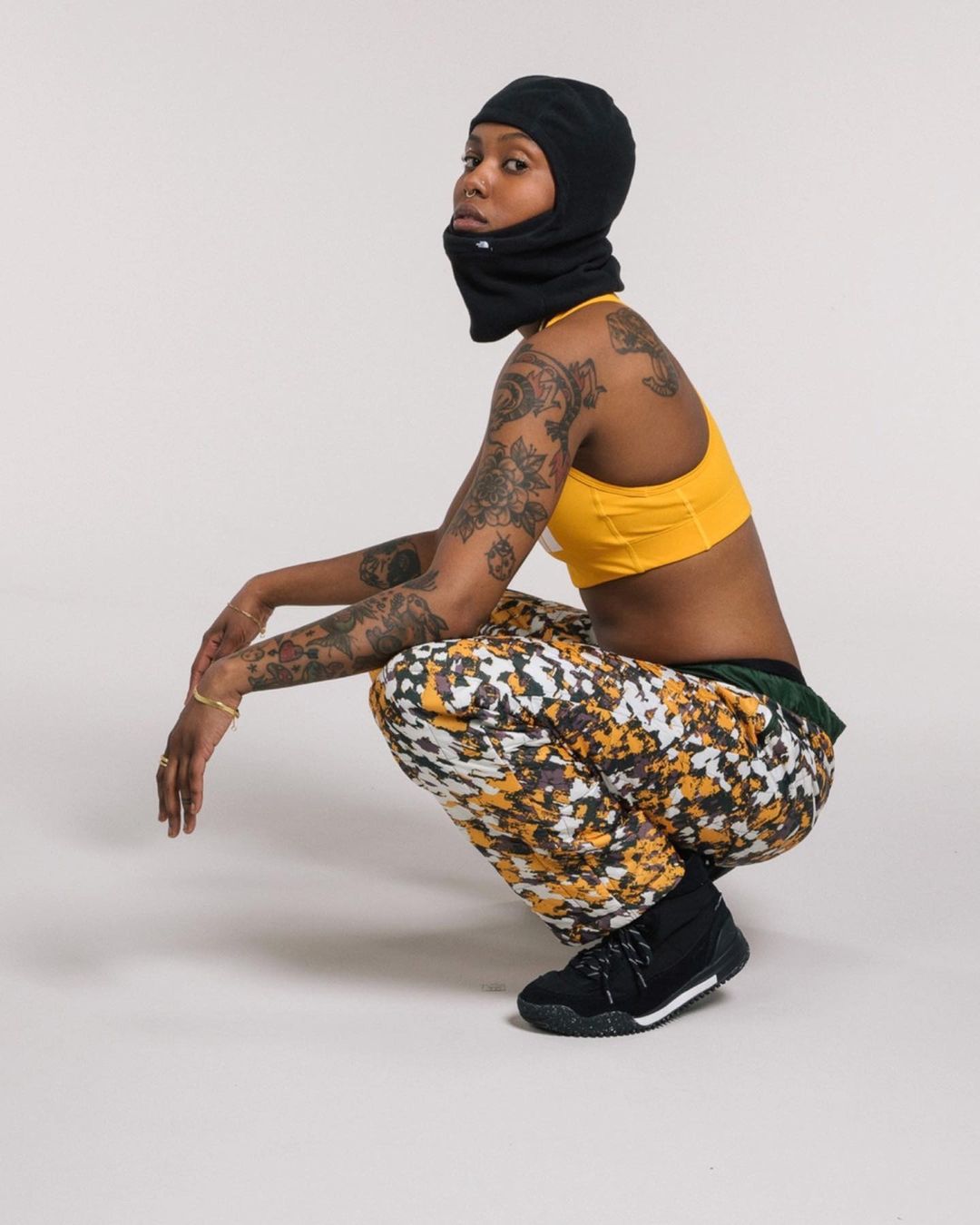 A woman in a black balaclava, yellow top, and patterned pants squats in a photo studio, showcasing fashion and personal style against a neutral backdrop.