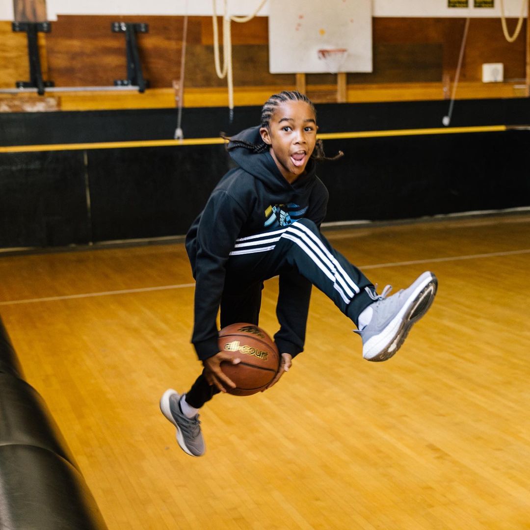A young boy in sportswear jumps indoors, dribbling a basketball on a wooden court, showcasing his athletic skills in an active shot.