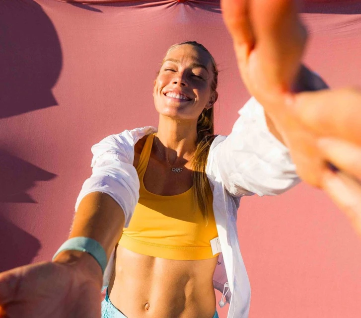 Smiling woman in athletic wear takes a selfie against a solid pink backdrop, showcasing fitness and active lifestyle.