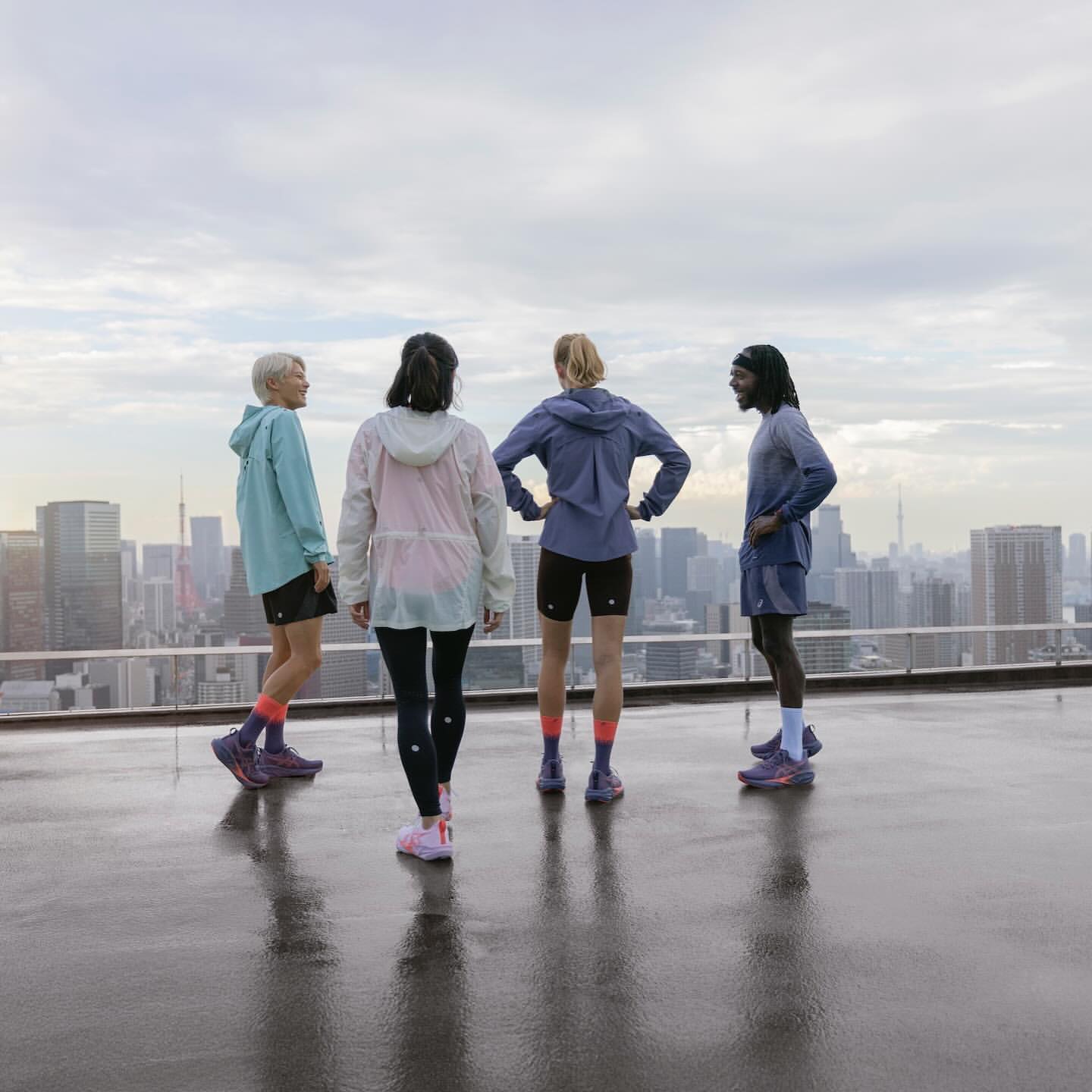 Four runners in athletic wear stand on a rooftop overlooking a cityscape under an overcast sky, showcasing fitness and urban athletics.