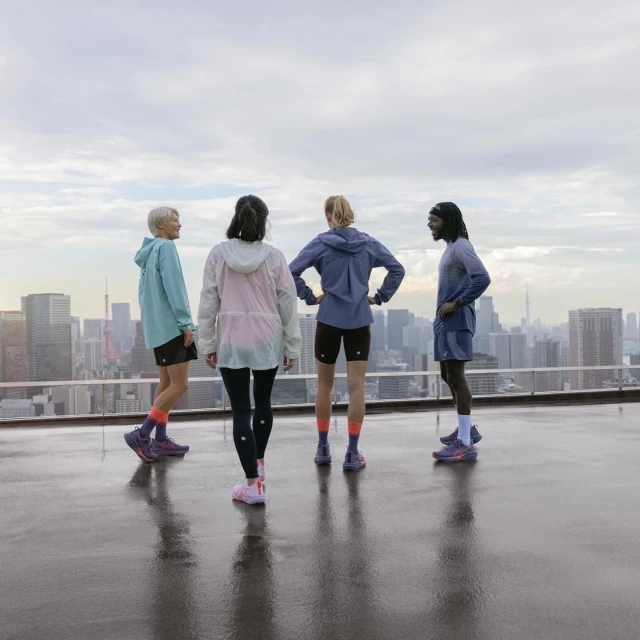 Four runners in athletic wear stand on a rooftop overlooking a cityscape under an overcast sky, showcasing fitness and urban athletics.
