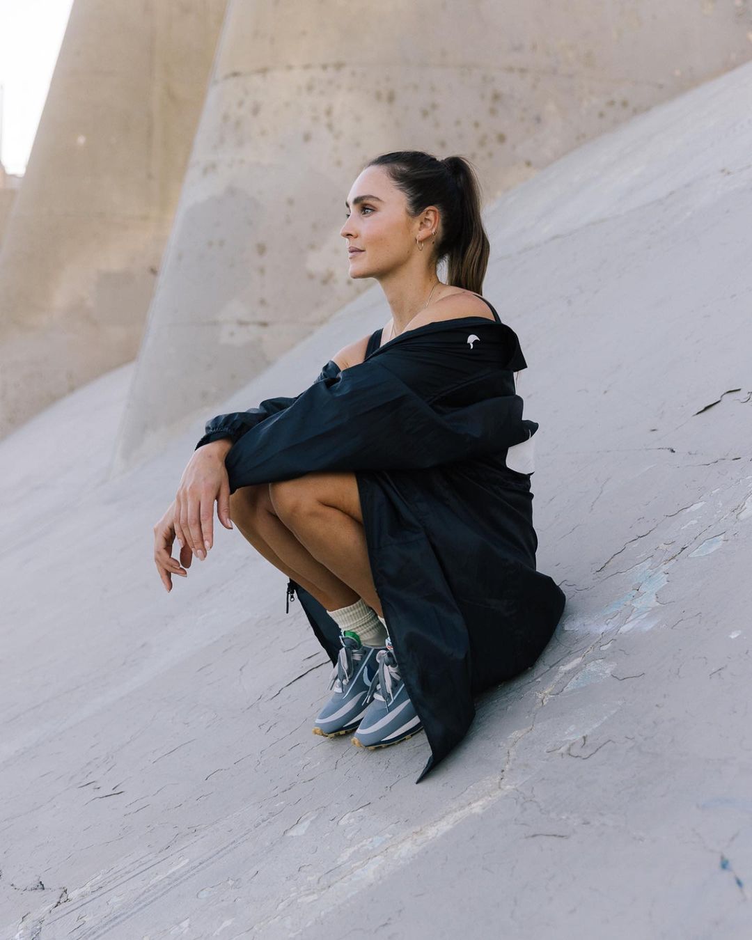 An adult woman in a dark jacket poses in a crouched position outdoors with a concrete structure as the backdrop in a lifestyle image.