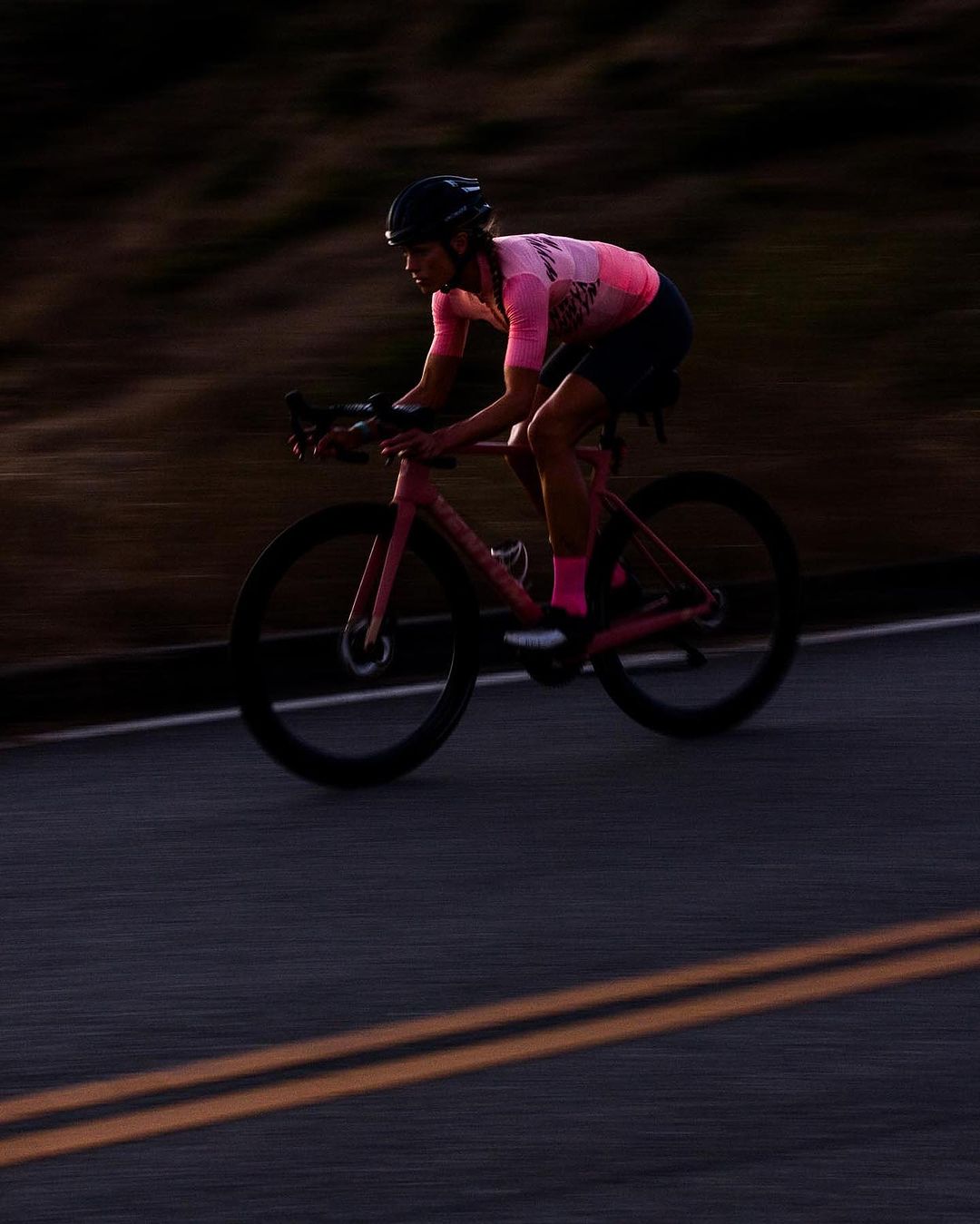 A person in a pink cycling jersey and black helmet rides a pink racing bicycle on an asphalt road in an outdoor setting.