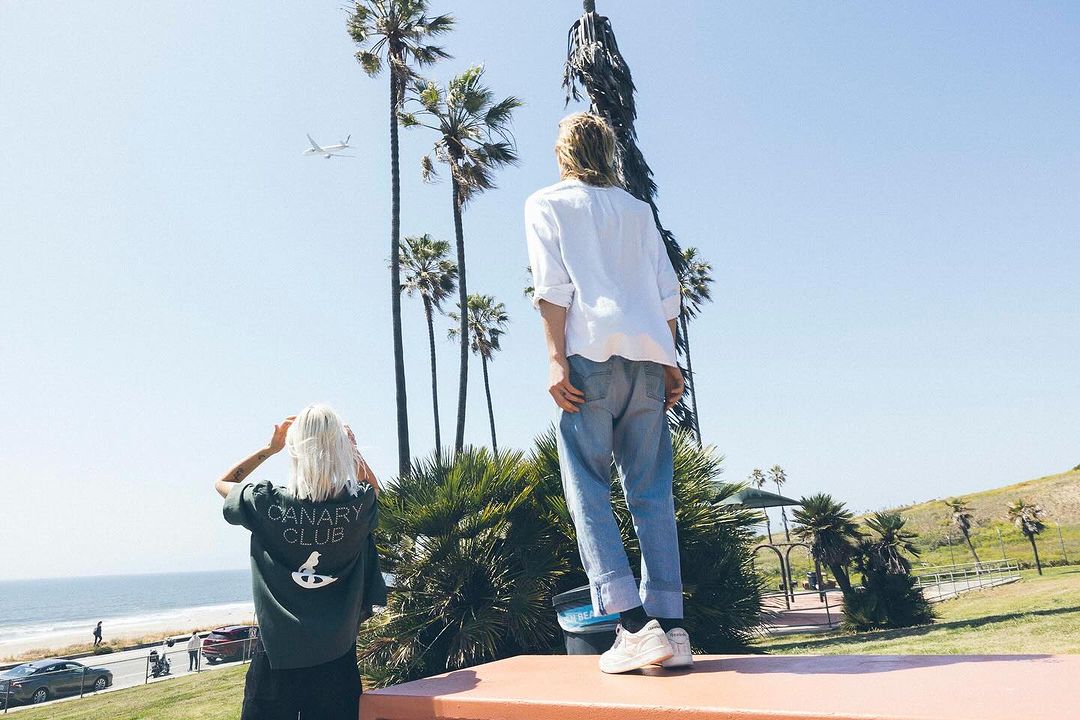 Two people stand on a platform overlooking the beach, palm trees, and ocean. An airplane is flying in the sky above the landscape.