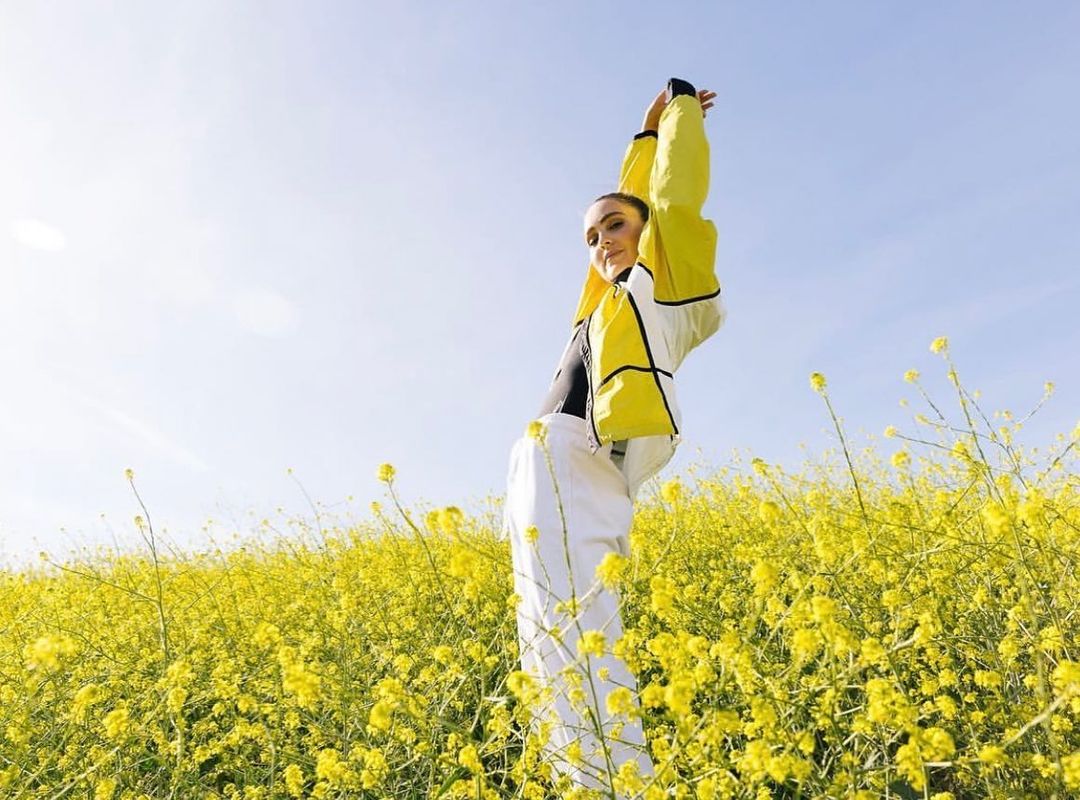 Young woman in yellow jacket and white pants poses in a field of yellow flowers under a clear sky, arms raised.