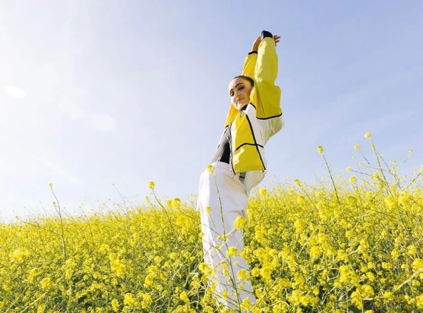 Young woman in yellow jacket and white pants poses in a field of yellow flowers under a clear sky, arms raised.