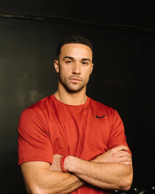 Portrait of an adult male with short dark hair, wearing a red t-shirt, with arms crossed in front of a dark grey background indoors.