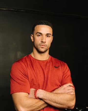 Portrait of an adult male with short dark hair, wearing a red t-shirt, with arms crossed in front of a dark grey background indoors.