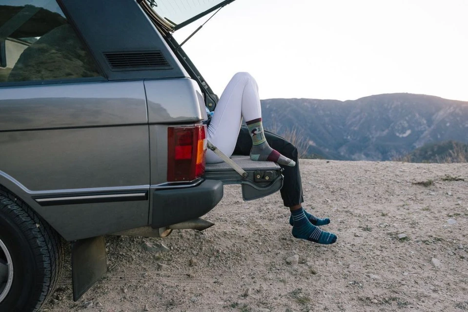 View of a person's legs in a vehicle trunk against a mountain backdrop, showcasing an outdoor, relaxed, and scenic setting with casual attire.