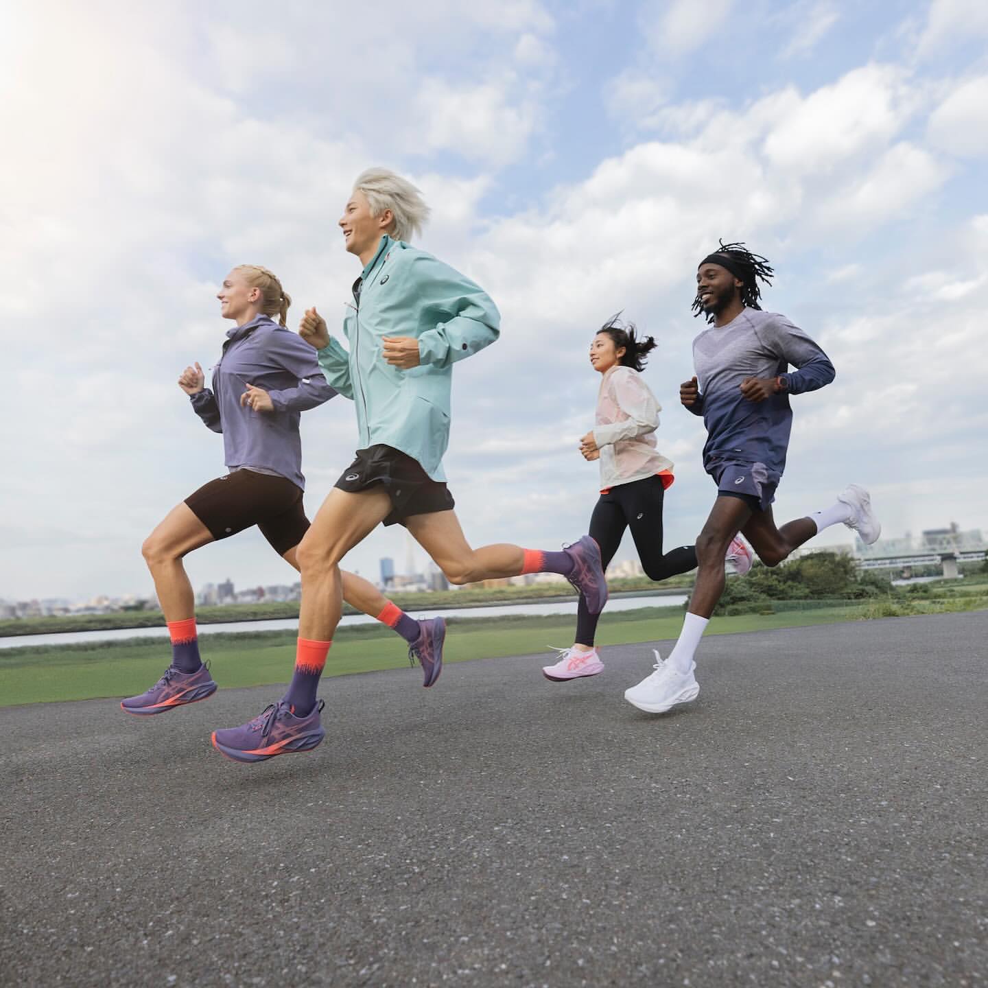 Four diverse athletes run at eye-level on a paved path. Runners wear jackets, shorts, running shoes, and long socks. Sky and cityscape backdrop.