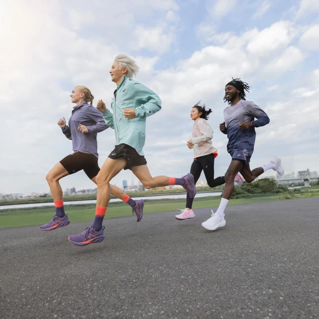 Four diverse athletes run at eye-level on a paved path. Runners wear jackets, shorts, running shoes, and long socks. Sky and cityscape backdrop.