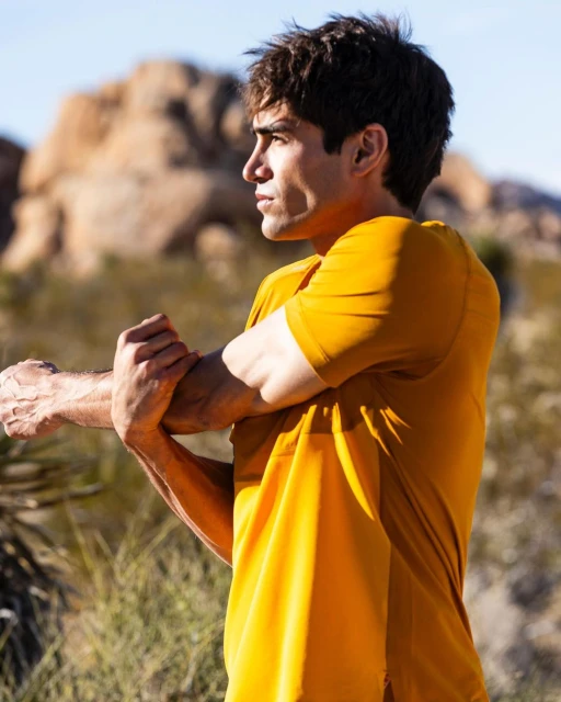 Athletic man in golden-yellow T-shirt stretching arms outdoors in a desert landscape with rock formations under bright sunlight.