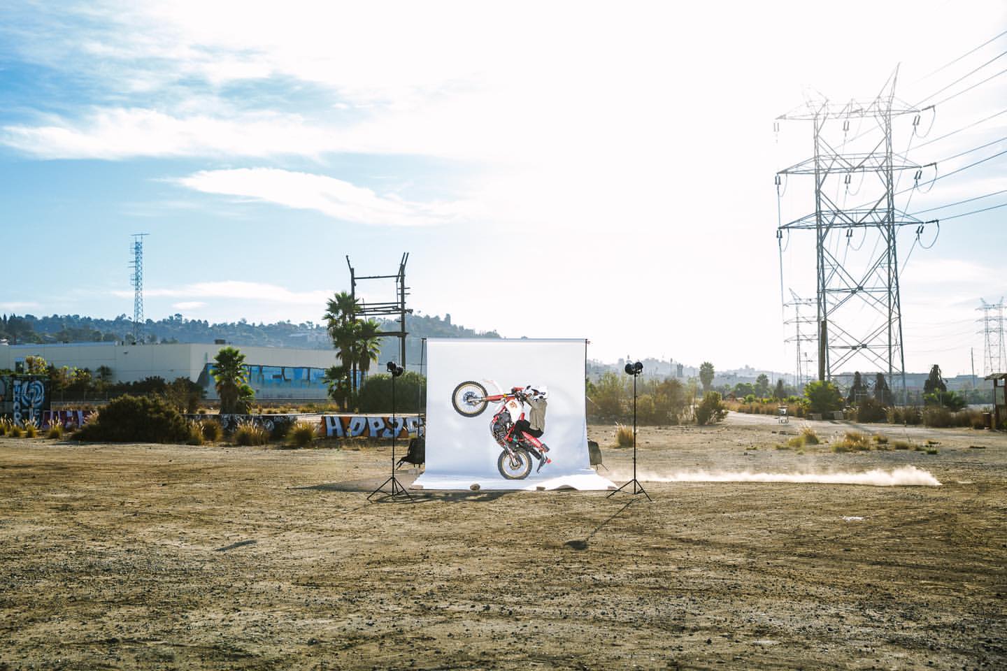 A person performs a wheelie on a red dirt bike in front of a white backdrop in an open field with power lines and a blue sky.