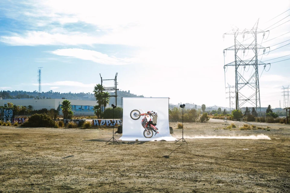A person performs a wheelie on a red dirt bike in front of a white backdrop in an open field with power lines and a blue sky.