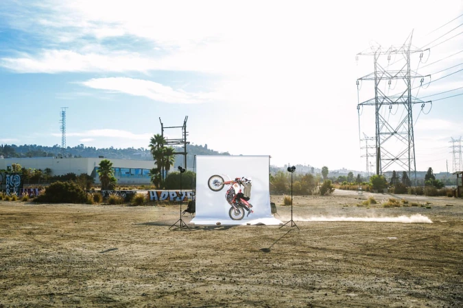A person performs a wheelie on a red dirt bike in front of a white backdrop in an open field with power lines and a blue sky.