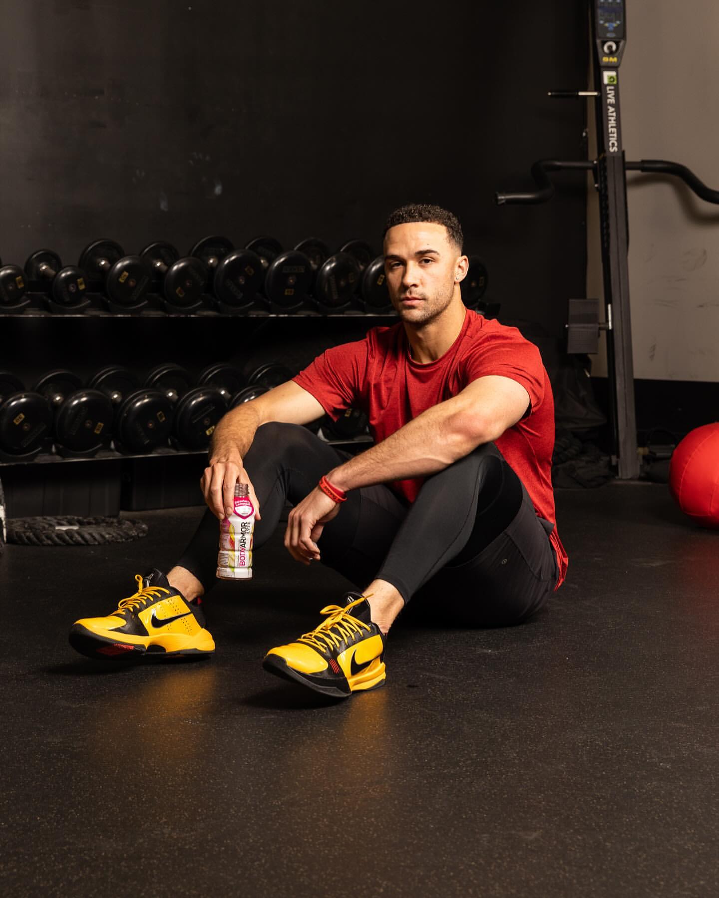 Athletic man sits on the floor in gym setting, wearing red shirt and yellow sneakers, holding a beverage, weights in background.