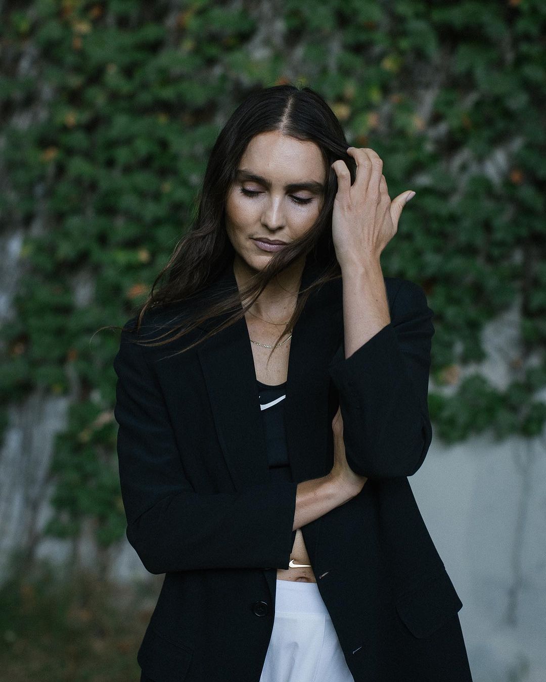 Portrait of a young woman outdoors wearing a black jacket and white skirt, hand in her hair, against a green leafy background.